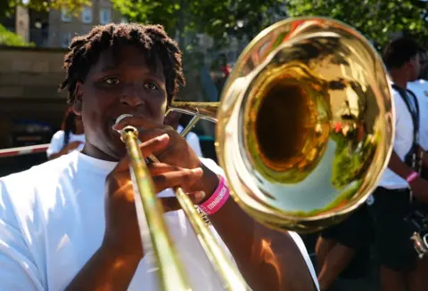Close-up of a musician playing a trombone outdoors in bright sunlight. The large brass bell of the instrument fills the foreground, with other band members visible behind.