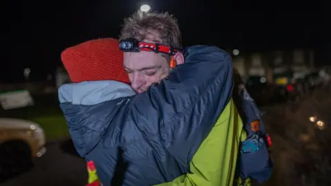 Wild Aperture Rhys Beddoe hugging his wife Hannah at the finish line of the Spine Race. Rhys is wearing a green waterproof and a red headtorch. His wife is wearing a navy waterproof and a red woolly hat. It is dark outside.