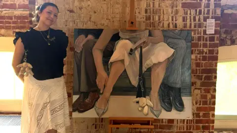 Contributed A woman stands next to a large piece of art. She is smiling and has dark hair that has been tied back. She is wearing a black blouse and a white skirt. The painting shows a woman sat on a tube who has spread her legs wide. She is encroaching on two men wearing suits sat beside her who have angled their legs away from her. The woman is wearing a white summer dress and has a black bag with a rabbit key chain attached to it that rests on the floor between her legs. She holds a book in one hand and her other hand is down toward her ankle. 