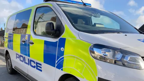 BBC The side of a police van. It is white, blue and yellow in colour. There is a blue light on top of the vehicle.