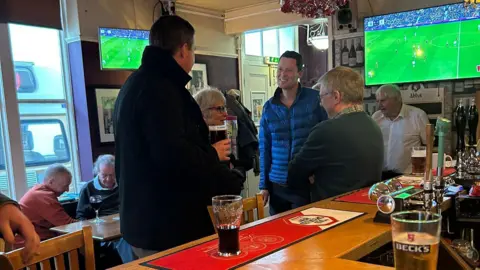 Submitted A group of people gather around a bar in a traditional British pub. There is a sports game on the TV, and people can be seen sat around a table in the background.