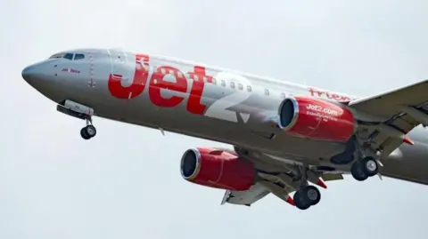 A Jet2 aeroplane flying against a pale grey sky. It is painted white and has Jet written in red and 2 written in white in large letters on its body.