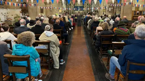 BBC/Oli Constable The picture shows the inside of a church, packed with people on every one of the 10 rows on either side. A woman stands at the end of an aisle in front of a long desk which has several people seated on it. Her back faces the crowd. Bunting is attached to the pillars of the church.