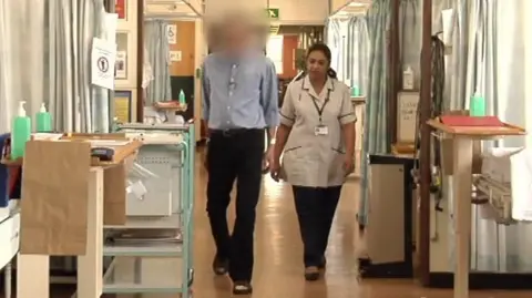 A man in a blue shirt and dark coloured trousers with his face blurred out walking next to a woman in a grey nurses uniform. They walk through a hospital corridor between beds with several wooden boards on metal plinths at the end of each bed and curtains visible.