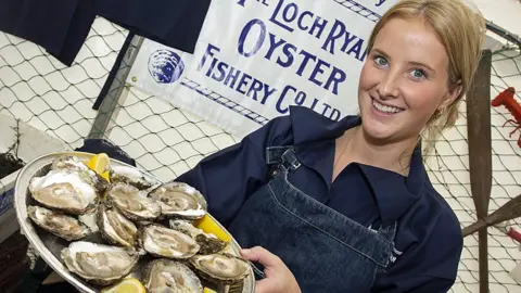 A young woman with blonde hair holds a tray of oysters
