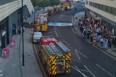 HIWFRS Large groups of people stand on the pavement on the opposite side of a line of fire engines outside a shopping centre.