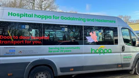 A bus with writing on the side. It reads: "Hospital hoppa for Godalming and Haslemere" and "Door to door hospital transport for you."
