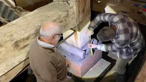 Caroline Peeke Two men are working on part of the frame within a mill. The frame is wooden with bricks at the bottom and one man is measuring part of it