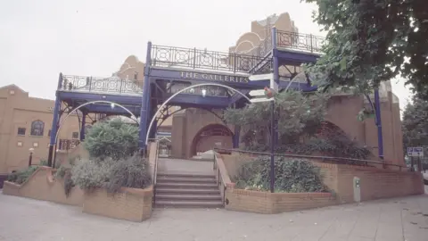 An image of one of the entrances to the Galleries as it currently looks. It has brick walls and steps running through blue railings, along with shrubbery. 'The Galleries' is seen on a blue arch over the entrance