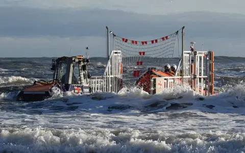 Helen Cowan An orange lifeboat is manoeuvred on to a trailer being pulled by a tractor which is submerged up to its cab in water.

