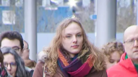 Dr Beth Upton - a trans woman with long hair - walks towards a hearing while wearing a maroon coat and multi coloured scarf. 