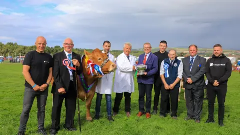 CALLUM STALEY/ CJS PHOTOGRAPHY Nine men smiling in a field, two wear white coats, and a cow with a number of rosettes stands among them.