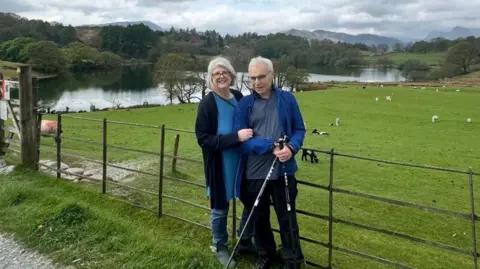 A woman and a man are standing alongside a metal fence with a field and large body of water on the other side. There are sheep in the field and in the distance is woodlands and further in the distance is a range of hills.