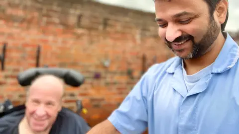 NIKKI FOX/BBC Dinesh Pushparajan in the courtyard of a care home in Bedford. He is to the right of frame looking down at a plant pot he is planting a plant in, while a resident is framed to the left looking at the camera in a wheelchair with a neck support. He is bald and is smiling. Dinesh has a beard and smartly shaven moustache.
