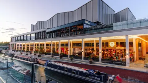 A new canal-side development features restaurants, a cinema and a rooftop bar. It is seen brightly lit at night with people gathered outside. Narrowboats are moored on either side of the canal below the development.