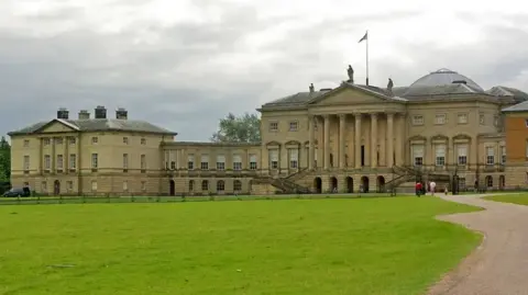 Kedleston Hall, which is a stately home, behind a patch of grass and grey skies
