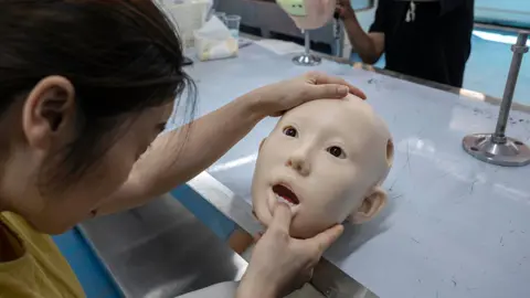 Getty Images A woman holding the head of a hairless human-like sex doll. She is inspecting its teeth.