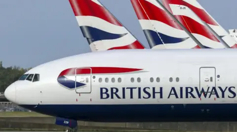 A white airliner with the words British Airways painted in blue, along with the blue and red logo above, is taxiing on an airport runway. There are four other British Airways planes in the background. 