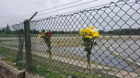 Henry Godfrey-Evans/BBC Floral bouquet that has been strapped to a metal fence. They are made of flowers that are different shades of yellow, orange, pink and red. A reservoir can be seen on the other side of the fence. It is a grey cloudy day.