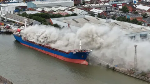 Dale Baxter/BBC An aerial view of a blue and red vessel with grey smoke billowing out from the deck. A number of industrial buildings are visible in the background.
