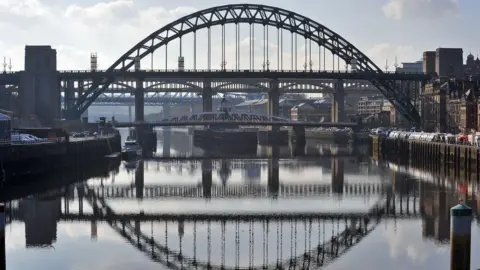 BBC The Tyne Bridge connecting Newcastgle and Gateshead. The bridge's reflection can be seen in the water. The Swing Bridge can be seen underneath the Tyne Bridge.