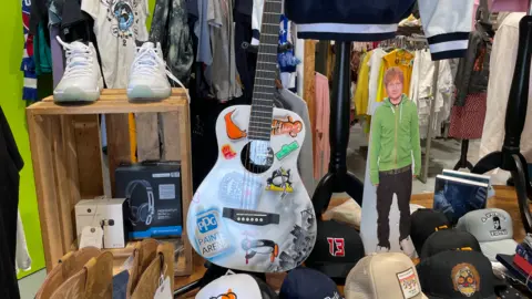An acoustic guitar surrounded by baseball cats positioned next to a wooden stand displaying a pair of trainers, headphones and earphones. An Ed Sheeran cut out and other T-shirts can be seen in the background.