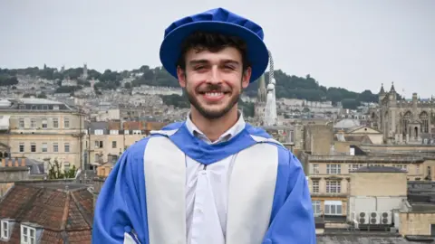 George Clarke is standing on a balcony overlooking the city of Bath. He is dressed in a graduation cap and gown after being given an Honorary Doctorate of Arts. The gown and cap are blue and silver. George has short dark hair and facial hair and is standing with his hands clasped in front of him, looking at the camera and smiling.