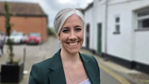 Daisy Cooper is standing in a street wearing a green jacket over a white top.