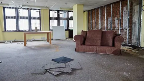 Hastings Commons A rundown office unit in Rock House containing a disused sofa, table and carpet mats in the foreground. Leftover bunting can be seen hanging from the curtain rails. 