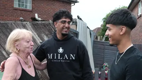 A small blonde woman wearing a black vest top and gold jewellery hugs a young man in a black hoodie with short curly black hair and a short beard another young man with black hair wearing a T-shirt stands beside them.