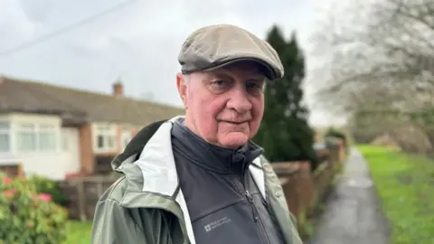 Tongham Resident Tom Scanlon looks straight at the camera. He is wearing a flat cap and green coat. Behind him is a country lane, with houses on one side. The sky is grey and the ground looks wet. His face is slightly pink.
