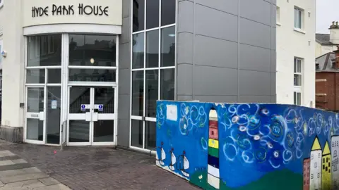 Front door of drug misuse charity Harbour's headquarters in Plymouth. A sign saying Hyde Park House is above the front doors, which are on the corner of a cream coloured building. A colourful mural has been painted on a wall next to the building.