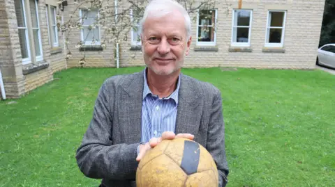 West Oxfordshire District Council Councillor Tim Sumner stands in a grass area, there is a building in the background with pale beige brickworks and a blossom tree, he stands in the middle of the of the image holding a yellow football with a black stripe. Councillor Sumner has white short hair and is looking directly at the camera, smiling and wearing a blue and white striped shirt with a grey tweed blazer over the top.