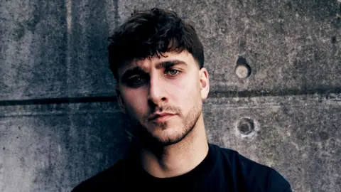Joe Magowan A man wearing a dark coloured top, standing in front of faded grey concrete bricks.