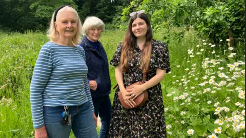 Three women stand side by side in a grassy area with white flowers next to them. It is a serene scene and they are smiling.