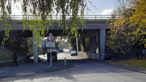 A small road leading to a residential area, with a concrete bridge over it.