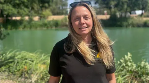 A woman with long blonde hair is standing in front of a fishing lake, wearing a black tee shirt and with sunglasses on her head.