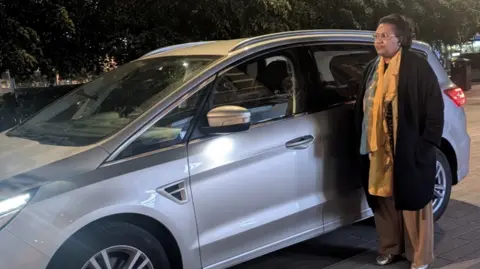 Lula Kinnaird has dark short hair and brown eyes. She is wearing a yellow outfit and black coat. She is standing next to her silver car which she uses for business.
