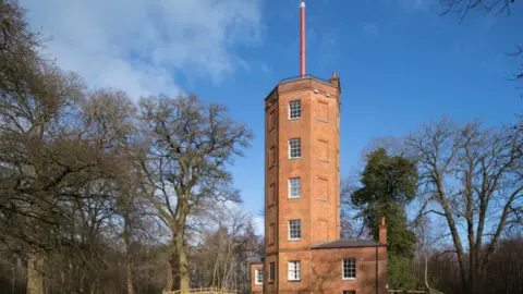 The Landmark Trust The picture shows a hexagonal tower made of brown bricks. There is a pole on the top of the building. There are rows of windows up and down the sides of the tower. Behind the structure there are green trees and a blue sky in the distance