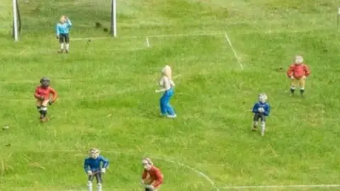 A close up of a football pitch with miniature model players. A figure of a topless blonde-haired woman can be seen running across the pitch. 