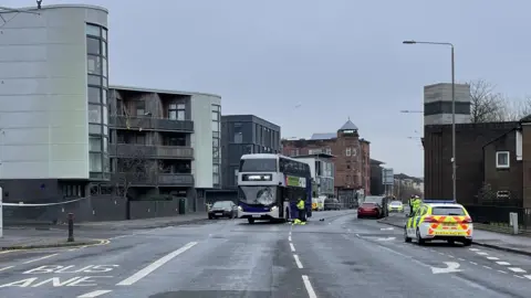 A street in Glasgow. A bus is stopped in the middle of the road, it has some damage to the windscreen. Police officers can be seen standing next to it.