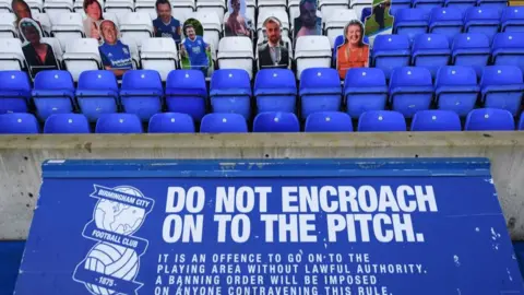 Getty Images Rows of blue and white seats at a football stadium. Most are empty but some are occupied by cardboard cutouts depicting fans. A sign with the Birmingham City FC logo on it warns: "Do not encroach on to the pitch."