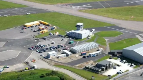 Newquay Airport from above. It has a long runway, a control tower and several buildings and structures. There are also several vehicles parked in the area, which is surrounded by green fields.