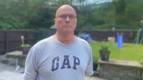 Simon Thake A cross stocky middle aged man with shaved hair wearing a grey t-shirt looks towards the camera