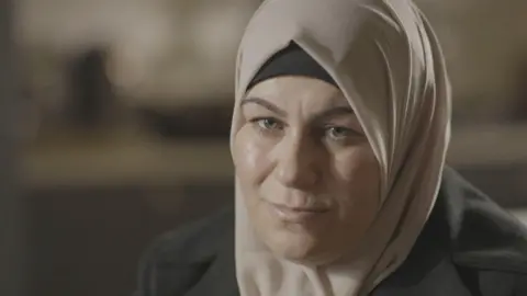 A woman dressed in a headscarf is sitting in a room looking to the side of the camera. The woman has green and hazel coloured eyes. Her headscarf is made of a fine silky material and is beige in colour. She is also wearing a dark coat. Behind her, the room is out of focus with nothing visible.
