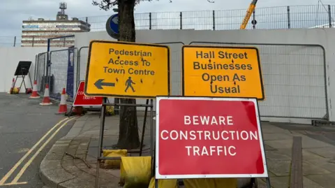 Three temporary road signs in front of a construction site. A yellow sign with black writing says: "Pedestrian access to the town centre with a black arrow and a silhouette of a person walking. A second yellow sign with black writing says: "Hall Street businesses open as usual". A red sign with white writing says: "Beware construction traffic".