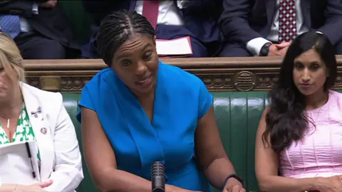 Kemi Badenoch at PMQs - she is wearing a blue dress leaning on the despatch box