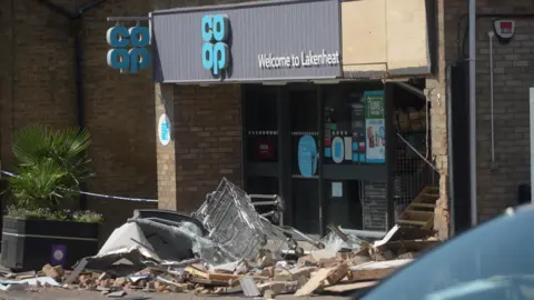 Knocked over trolleys lie on top of wooden and brick debris outside a Co-op. The wall is damaged and there is a hole in the front of the shop.