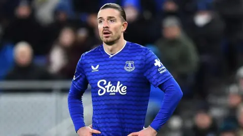 Reuters Everton's Jack Grealish stood on a fotball pitch wearing a blue Everton FC shirt with his hands on his hips