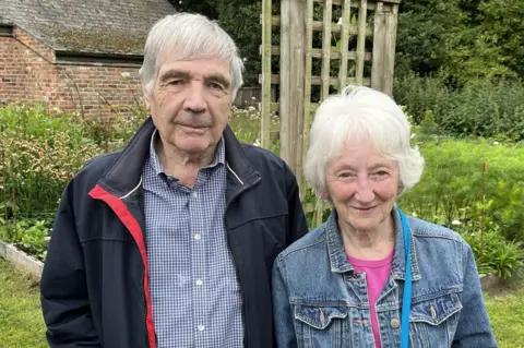 A man with grey/white hair brushed forwards and wearing a blue and white check shirt and dark navy jacket with a red lining stands next to a woman with short white, wavy hair, she is wearing a denim jacket over a pink t-shirt, with a blue strap across her shoulder. They are standing in a garden with a flower bed, trellis and old stone wall in the background